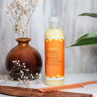 Bottle of 'HEADS UP' shampoo on a surface with a vase, comb, and flowers in the background.