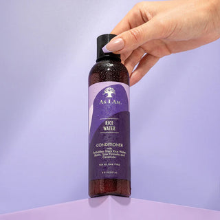 Hand holding a bottle of As I Am Rice Water Conditioner against a light purple background