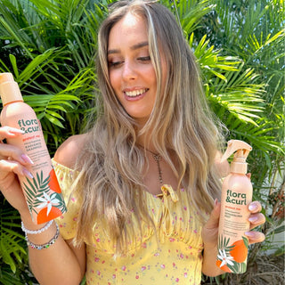 Woman holding two bottles of 'flora & curl' products in front of green foliage