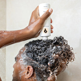 Person applying a white bottle of shampoo to their curly hair with a neutral background. applying Bottle of Bounce Curl Gentle Clarifying Shampoo on his hair
