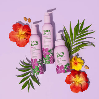Three 'flora & curl' hair care products with tropical flowers and leaves on a light purple background