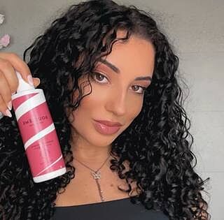 Woman holding a pink and white spray bottle with curly hair against a neutral background