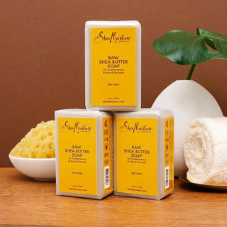 SheaMoisture Raw Shea Butter Soap packaging on a wooden surface with a plant and bowl in the background.