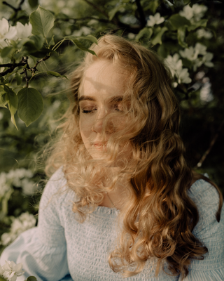 Woman with long wavy blonde hair sitting among green foliage and white flowers