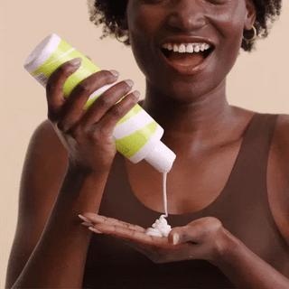 Woman applying a white cream from a green and white container on her palm against a beige background